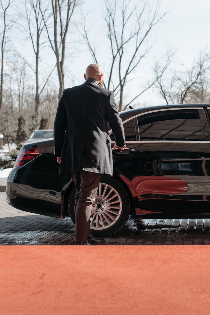 Bald man in a black suit approaches a luxury black car on a sunny day.